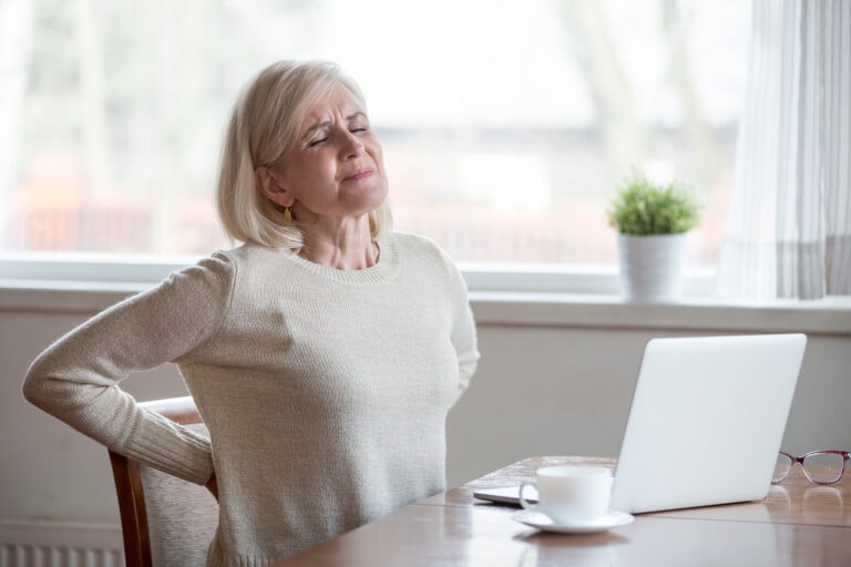 Woman experiencing lower back pain while sitting at a desk and working on a laptop